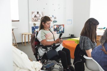 ve workshopové místností sedí mladá tmavovlasá žena na vozíku a čte z tabletu, okolo sedí další posluchači.